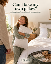 A young girl in green floral pyjamas hugs a white pillow and looks up at her parent, smiling. An open bag on the floor holds a teddy bear and folded clothes. A neatly made bed with white and sage bedding is visible behind her.