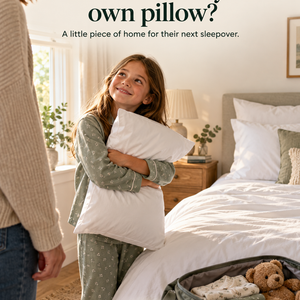 A young girl in green floral pyjamas hugs a white pillow and looks up at her parent, smiling. An open bag on the floor holds a teddy bear and folded clothes. A neatly made bed with white and sage bedding is visible behind her.